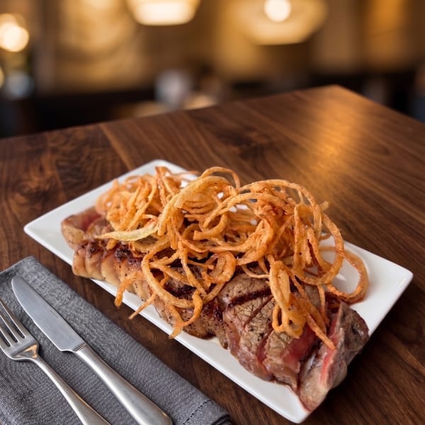 Sliced steak topped with crispy fried onion rings on a white platter, with cutlery on a dark wooden table.