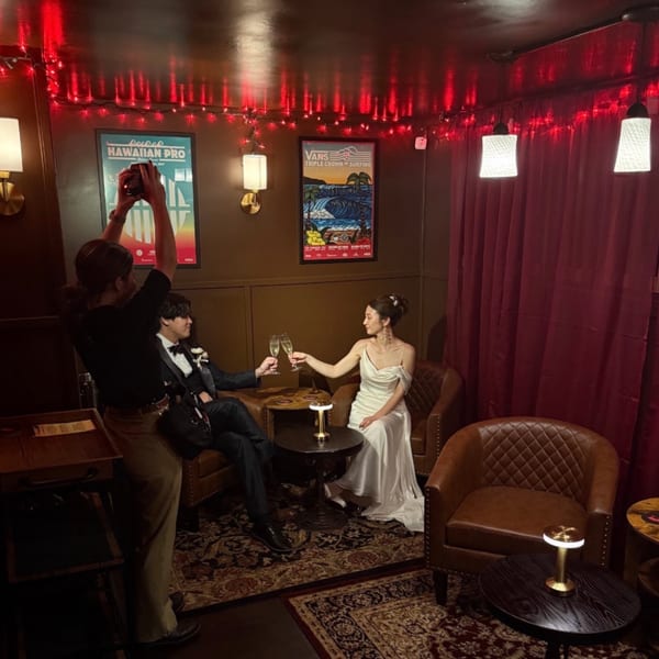 A couple in formal wear toasts with champagne in a dimly lit room with red string lights and surfing posters.