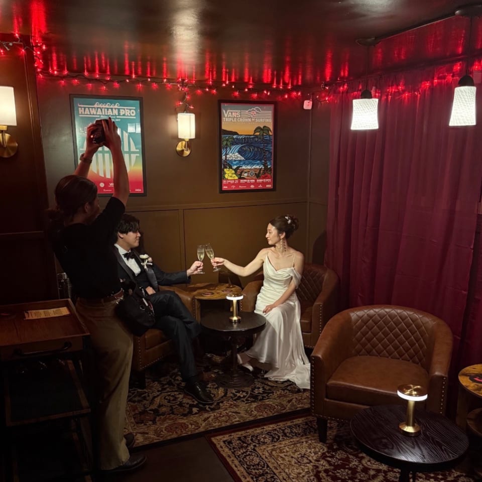 A couple in formal wear toasts with champagne in a dimly lit room with red string lights and surfing posters.
