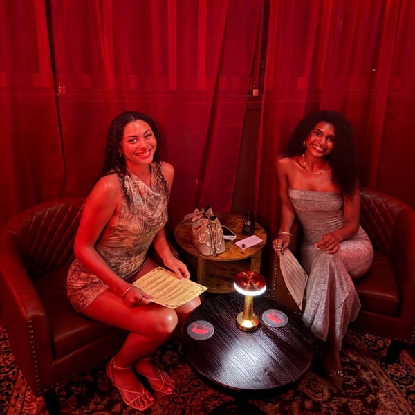 Two women smiling in red-lit armchairs, holding menus beside a small table with a lamp.