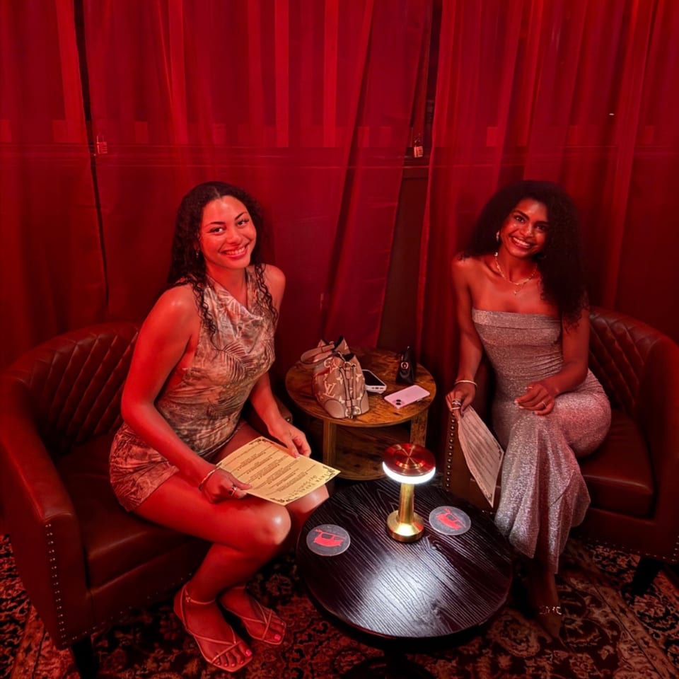 Two women smiling in red-lit armchairs, holding menus beside a small table with a lamp.