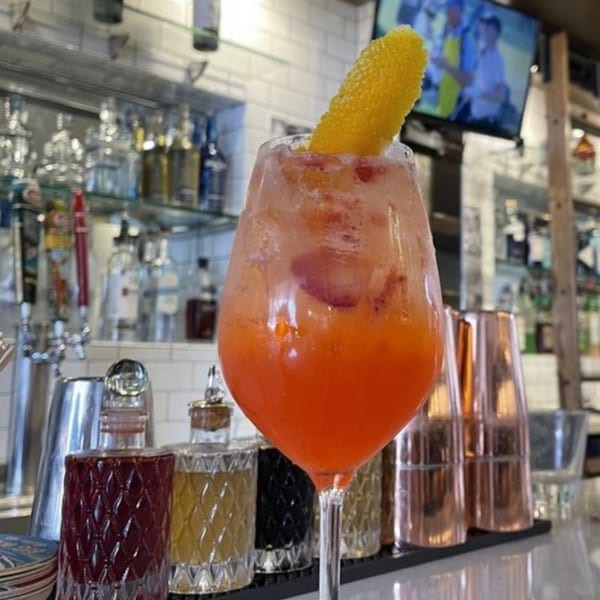 A vibrant orange cocktail with a lemon peel garnish sits on a bar with bottles and a TV in the background.