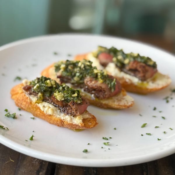 Three crostini appetizers with meat, creamy spread, and green relish on a white plate.