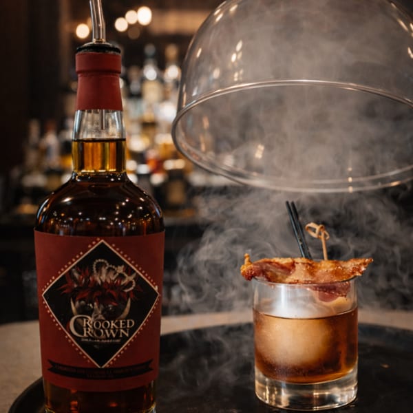 Smoked cocktail with bacon garnish next to a bottle of Crooked Crown whiskey at The Surfing Pig.