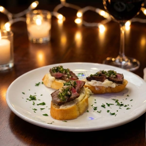 Three steak crostini with chimichurri on a white plate, with candles and wine in the background.