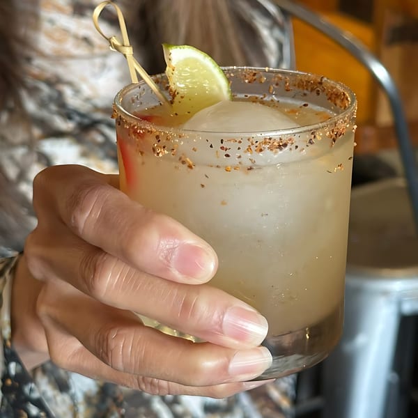 A hand holds a cocktail with a lime wedge, large ice cube, and a spiced rim.