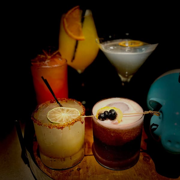 Five colorful cocktails, some garnished with fruit, on a dark wooden surface at The Surfing Pig.