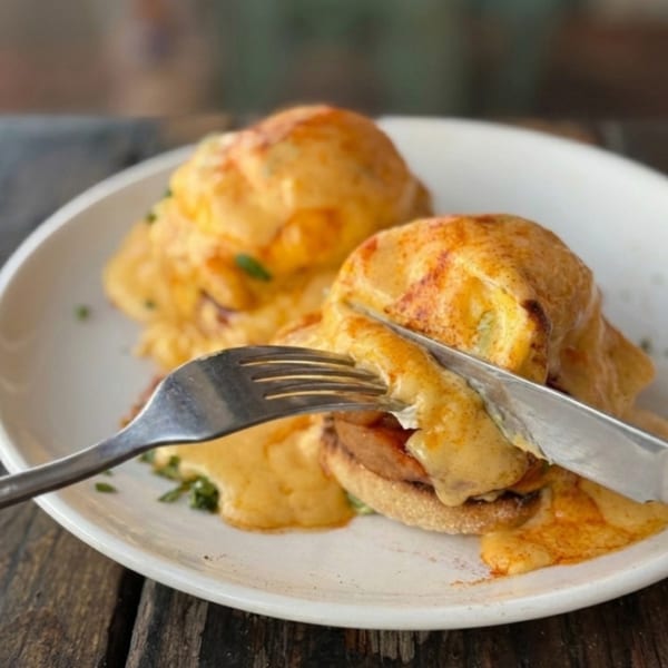Two eggs benedict with hollandaise sauce on a white plate, being cut with a knife and fork.