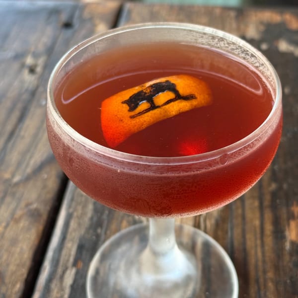 A red cocktail in a coupe glass, garnished with an orange peel featuring a branded pig.