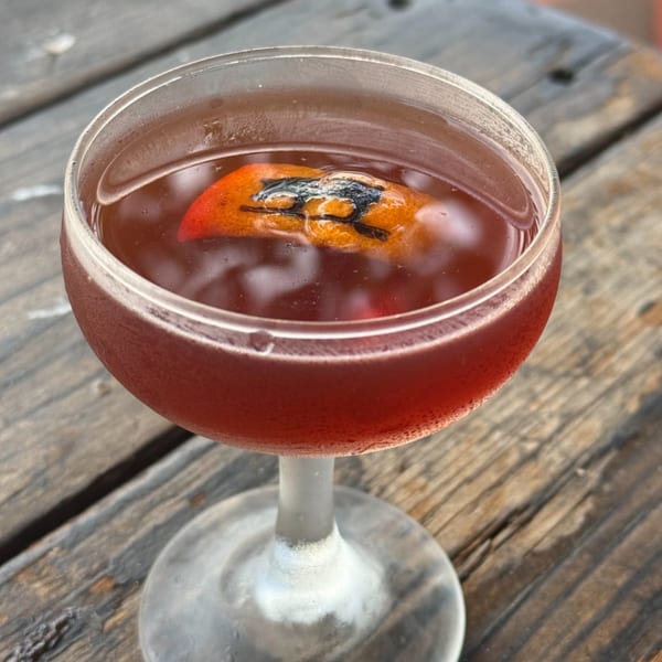 A red cocktail in a coupe glass, garnished with an orange peel featuring The Surfing Pig logo.