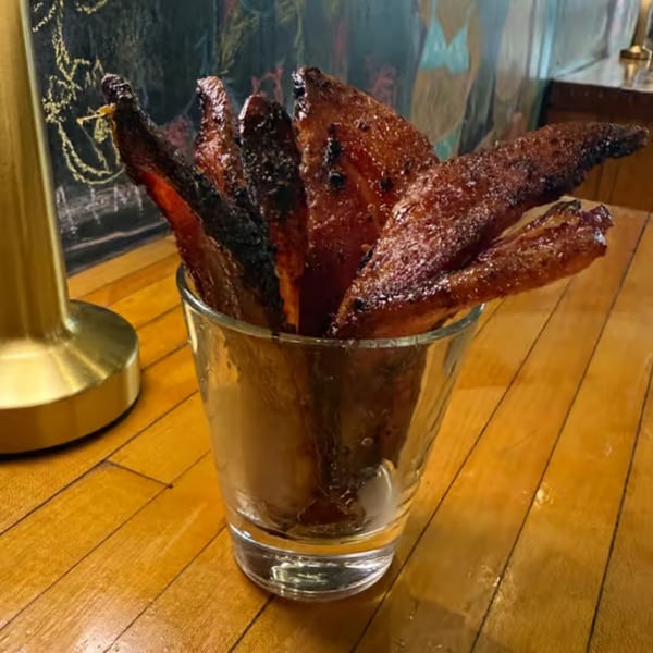 Crispy, dark brown bacon strips, some charred, standing upright in a clear glass on a wooden table.