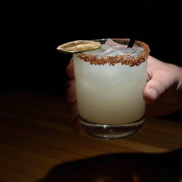 A hand holds a margarita with a chili-lime rim and a dried lime slice, against a dark background.
