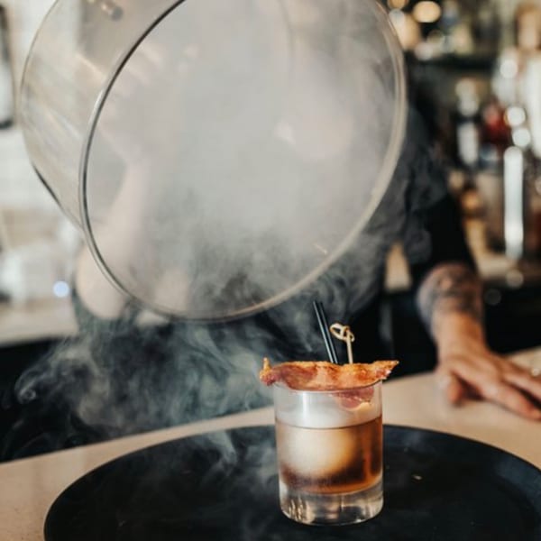 A smoked cocktail with a bacon garnish, served on a black tray at The Surfing Pig.